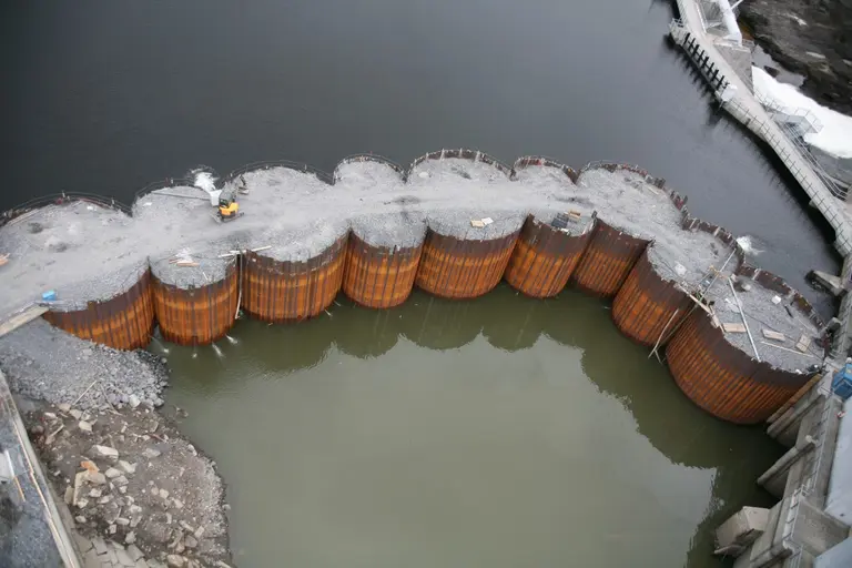 An aerial view shows a curved sheet pile cofferdam forming a temporary enclosure along a waterfront construction site.