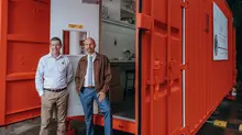 Two men standing beside an open red container office with a visible interior workspace.