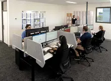 Office staff working at computers in a modern workspace while a colleague updates a planning board on the wall.