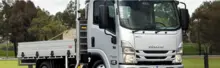 A white Isuzu flatbed truck parked outdoors, carrying a load of gravel, with trees and grass in the background.